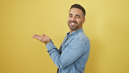 Sticker - Cheerful young hispanic man in denim shirt enthusiastically presenting ad, gesturing excitedly towards open copy space on yellow isolated background.