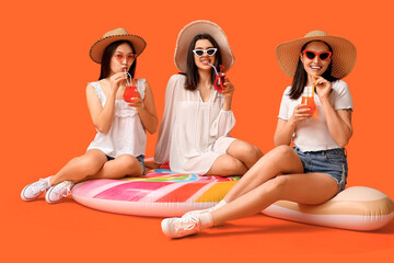 Wall Mural - Beautiful young women with glasses of cocktail sitting on inflatable mattress against orange background. Summer party