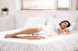 © Pixel-Shot - Young woman in pajamas with soft orthopedic pillow for legs on cozy bed at home