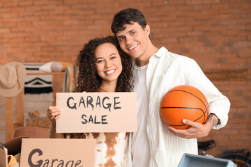 Canvas Print - Young couple holding cardboard with text YARD SALE and ball in room of unwanted stuff