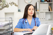 © JackF - Portrait of female doctor in surgical scrubs sitting at working table in her office and using laptop.