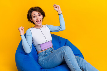 Poster - Portrait of overjoyed nice girl sit comfy bag raise fists shout yeah luck isolated on yellow color background