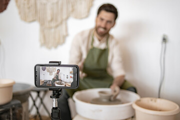 Young male potter sculpts a clay jug using a potter's wheel while recording a video blog with a smartphone.