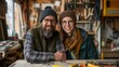 © Stone Story - Happy couple wearing winter hats and glasses in a cozy workshop filled with woodworking tools, smiling and leaning on a workbench.