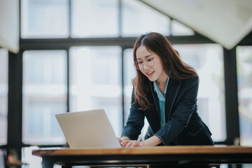 Wall Mural - Young Asian professional business woman office worker analyst sitting at desk working on laptop thinking on project plan, analyzing marketing or financial data online, watching elearning webinar.