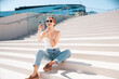© halayalex - Beautiful smiling model in sunglasses. Female dressed in summer hipster white T-shirt and jeans. Posing near white wall in the street. Funny and positive woman having fun outdoors, sits at stairs