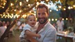 © Irina Ukrainets - Smiling father holding baby at outdoor evening event. Warm, welcoming atmosphere with string lights hanging. Perfect for family, celebration, or lifestyle themes, . AI