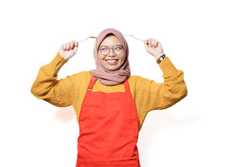 Wall Mural - Smilling young Asian Muslim woman wearing red apron holding spoon and fork isolated on white background