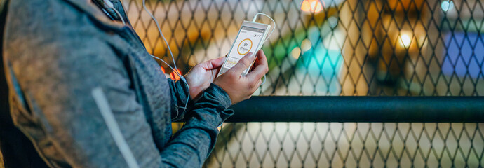 Banner of woman runner holding mobile phone with heart rate monitor on screen after training on city at night. Unrecognizable female athlete holding cellphone with activity summary on tracker app.