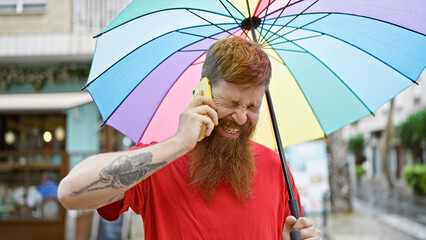 Poster - Cheerful young redhead man, confidently enjoying a funny chat on his smartphone, looking up smiling from under his umbrella on a city street.