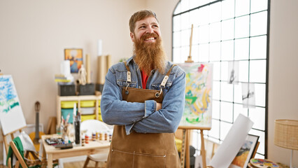 Sticker - Bearded handsome redhead man, young artist, stands confidently in art studio with arms crossed, smiling brightly while enjoying his painting class