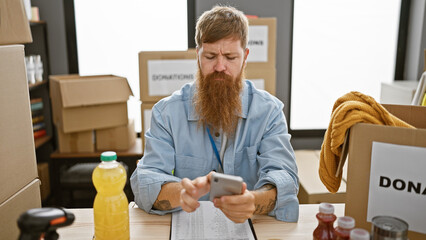 Sticker - Handsome, redhead volunteer sitting at table, focused on smartphone, making online donations in charity center