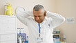 © Krakenimages.com - A middle-aged hispanic man in lab attire expresses stress or headache in a clinical hospital setting.