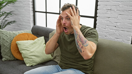 Wall Mural - Shocked hispanic man with tattoos expressing surprise indoors on a comfy sofa.