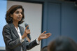 © ramona - an indian woman giving speech in an university aula with students, speaking into microphone with hand outstretched pointing towards camera, wearing grey suit jacket and white blouse underneath