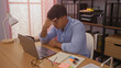 © Krakenimages.com - A young hispanic man looking stressed sits at a desk in an office, holding his forehead while working on a laptop surrounded by files, notebooks, and office supplies.