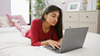 © Krakenimages.com - A focused south asian woman works on her laptop while lying on a white bed in a cozy bedroom.