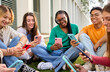 © CarlosBarquero - Group of Caucasian young people sitting on the grass looking smiling videos on cell phone outdoor. Generation z students friends having fun standing watching mobile together on university campus
