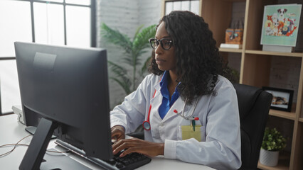 Wall Mural - A young african american woman doctor with curly hair works on a computer in a clinic's indoor workplace wearing a white coat and a stethoscope, focusing on her medical tasks.