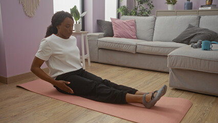 A young african american woman with curly hair is sitting on a mat, exercising in a cozy living room of her home, showcasing a calm and relaxed indoor setting.