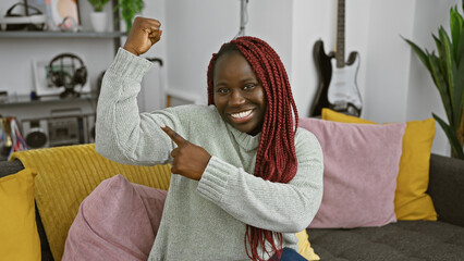 Wall Mural - A smiling african american woman with braids flexing in a cozy, modern living room interior.