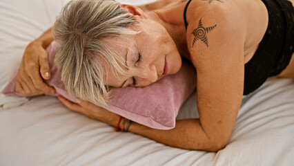 Mature woman with grey hair and tattoo rests on bed in a serene indoor bedroom setting.