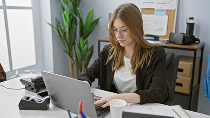 A focused caucasian woman working on a laptop in a modern office setting, displaying professionalism and concentration.