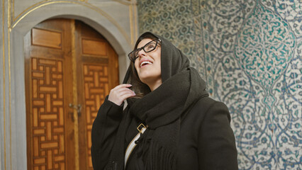 Wall Mural - A smiling young woman with glasses wearing a headscarf stands in front of traditional turkish tiles at istanbul's topkapi palace.