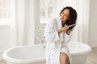 © Home-stock - Haircare. African american woman brushing hair with comb, detangling split ends and smiling, sitting on bathtub in white bathrobe. Beauty routine concept