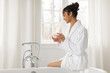 © Home-stock - Young pretty black woman in bathrobe holding cosmetic cream jar, sitting on bathtub in modern bathroom interior, free space