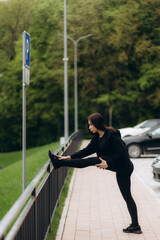 Wall Mural - Athletic fitness woman stretching her leg on the fence outdoors. Sporty woman warming up before outdoor workout in the park.