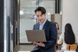 © amnaj - Confident businessman working on a laptop in a modern office environment with colleagues in the background. Professional and focused.