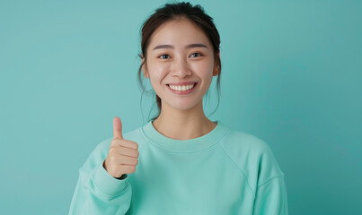 Wall Mural - Young asian woman in a mint sweatshirt smiling with healthy teeth showing thumb up at copy space expressing wow emotion standing isolated on mint background
