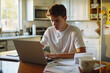 © GustavsMD - Young man working on laptop at kitchen table with documents and coffee cup. Home study and remote learning concept. Indoor candid photography
