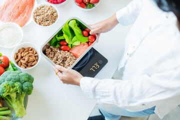 Healthy diet plan for weight loss, daily ready meal menu. Close up Woman weighing lunch box cooked in advance,ready to eat on kitchen scale. Balanced portion with healthy dish. Pre-cooking concept