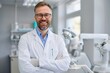 © Anton Gvozdikov - A smiling male dentist with eyeglasses standing confidently with crossed arms in a modern, well-equipped dental clinic.