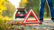© Irina Ukrainets - Emergency roadside triangle warning sign. Car breakdown safety equipment on a rural road. Autumn leaves scattered around. Vehicle and person in background. AI