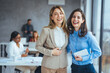 © Dragana Gordic - Two cheerful Caucasian businesswomen share a laugh, one holding a tablet, in a well-lit office while diverse colleagues focus on their tasks in the background.