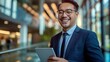 © nopommajun - Asian professional male smiles while holding a tablet standing outside an office