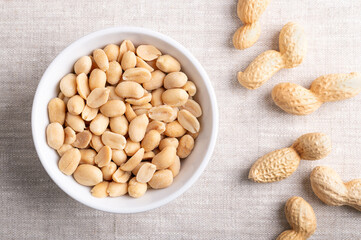 Wall Mural - Roasted and salted peanuts in a white bowl on linen fabric. Ready-to-eat snack food, made from fruits of Arachis hypogaea, also known as groundnut, goober, pindar or monkey nut. Close-up from above.