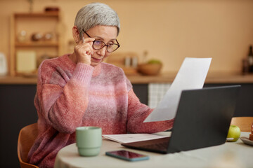 Portrait of Asian senior woman wearing eyeglasses reading document and doing taxes at home copy space