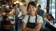 © MD Media - Happy woman restaurant owner standing in front of her cafe with arms crossed, smiling at the camera while customers sitting in the background, female business entrepreneur wearing an apron