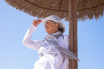 Wall Mural - Smiling senior woman dressed in white standing outdoors under a straw umbrella in a sunny day Elderly lady relaxes enjoying freedom in retirement or summer vacation