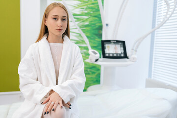 Wall Mural - Front view of elegant young woman patient in white bath robe sitting on medical couch in modern cosmetology clinic while waiting beautician specialist, looking at camera. Concept of spa treatment.