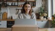 © Supranee - Happy young woman unboxing a package in a cozy kitchen, smiling at camera while opening her delivery, surrounded by plants and kitchen items.