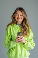 Wall Mural - 25 years old beautiful cute woman in a lime coloured sweatshirt on a grey background smiling, professional photo no letters holds the smartphone 