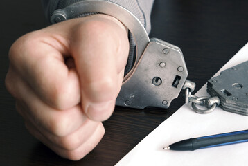 A close-up of a handcuffed man's hand clenched into a fist on a table. The picture shows a scene of frustration, anger and helplessness.