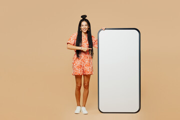 Wall Mural - Full body young Latin woman she wearing orange dress casual clothes point finger on big huge blank screen mobile cell phone smartphone with area isolated on plain beige background. Lifestyle concept.