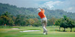 © alenagurenchuk - A senior golfer in a white hat and orange shirt swings his golf club on a green golf course with lush trees and a cloudy sky