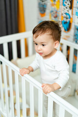 Poster - A baby is standing on a crib rail, looking at the camera. The crib is white and has a colorful design on the wall behind the baby